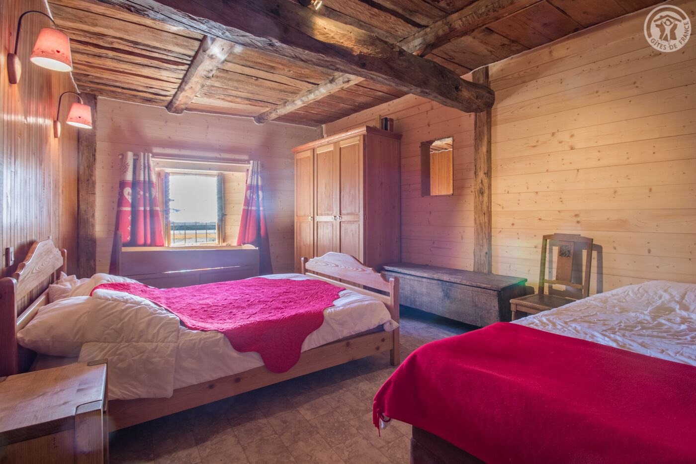 A cozy cabin room with two beds, wooden walls, a window with curtains, and a wooden ceiling.