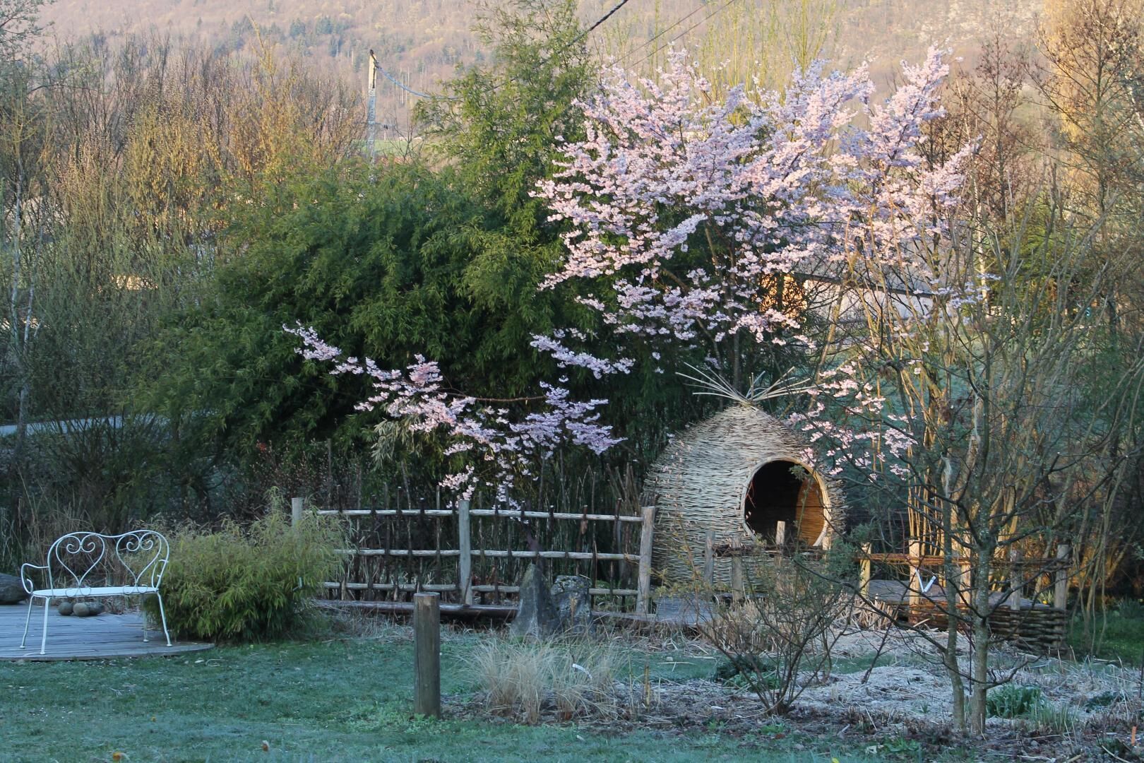 A rustic outdoor setting with a wooden structure resembling a hut, surrounded by blooming trees and a metal bench.