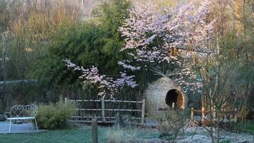 A rustic outdoor setting with a wooden structure resembling a hut, surrounded by blooming trees and a metal bench.