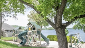 Ein Spielplatz mit Rutsche und Schaukeln, ein Haus mit einem begrünten Dach und ein See im Hintergrund.
