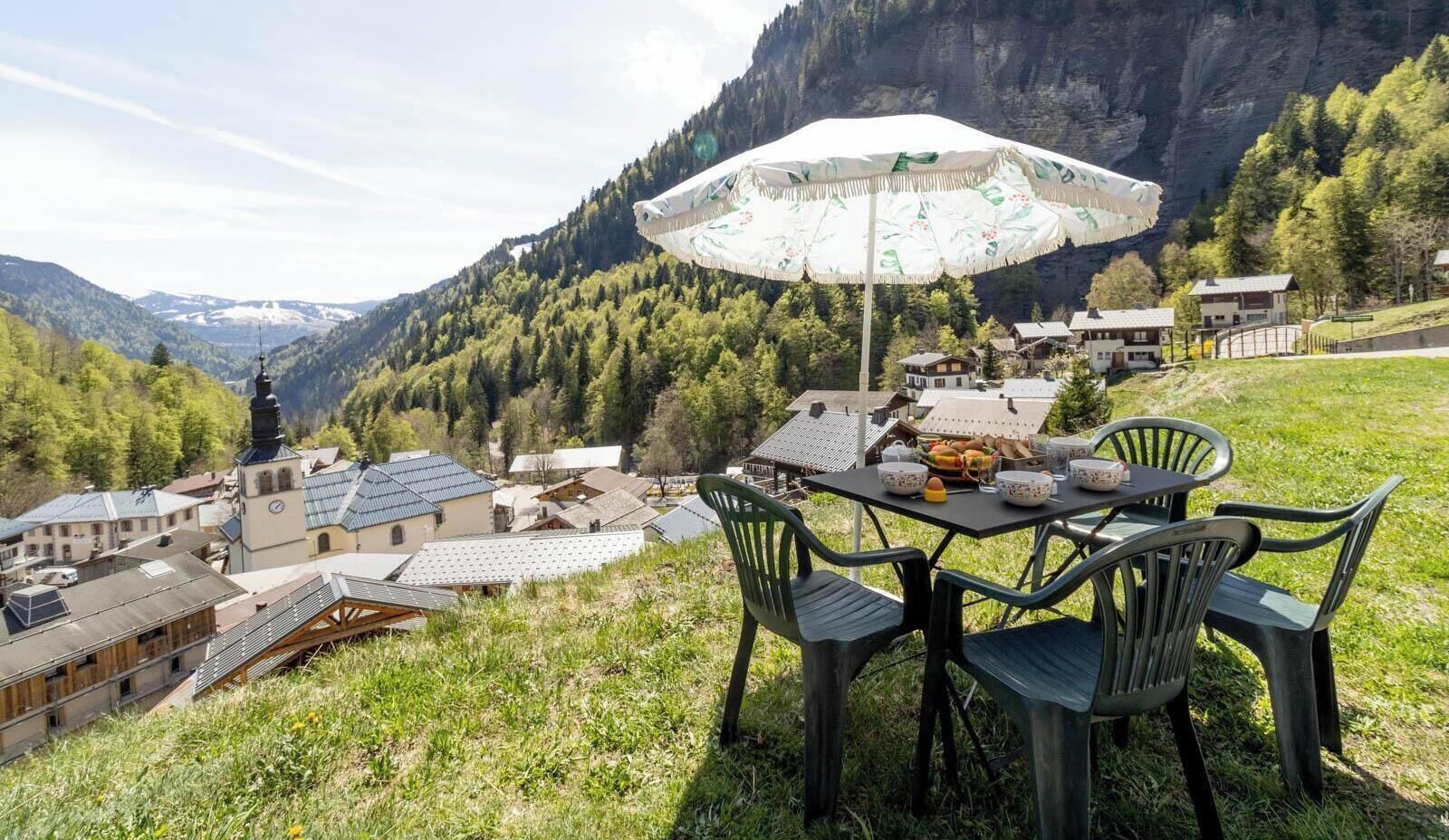 Ein Tisch mit Stühlen im Freien, mit Blick auf ein Dorf mit traditionellen Gebäuden und einer Bergkulisse.