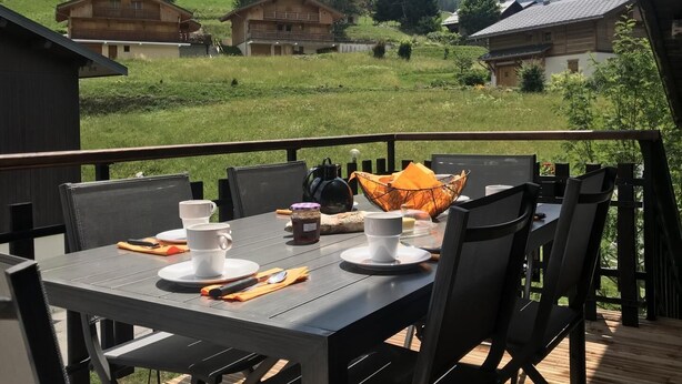 A dining table set with white cups, a bowl, and a spoon on a wooden deck with a view of green hills and houses.