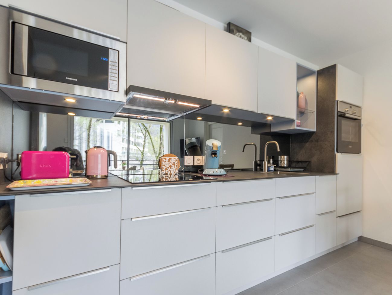 A modern kitchen with white cabinetry, a stainless steel range hood, a microwave, a kettle, and a coffee maker.