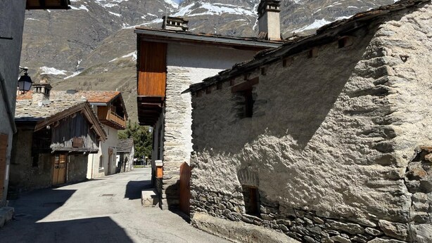 Eine enge Gasse mit Steinhäusern, eine Holzhütte mit Satteldach und verschneite Berge im Hintergrund.