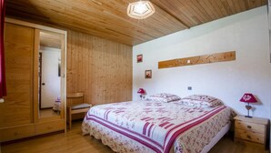 A bedroom with a wooden ceiling, a large bed with floral bedding, a wooden wardrobe, a mirror, and a wall-mounted artwork. - LA CHARMETTE - LA ROSIERE MONTVALEZAN (LA ROSIERE MONTVALEZAN)
