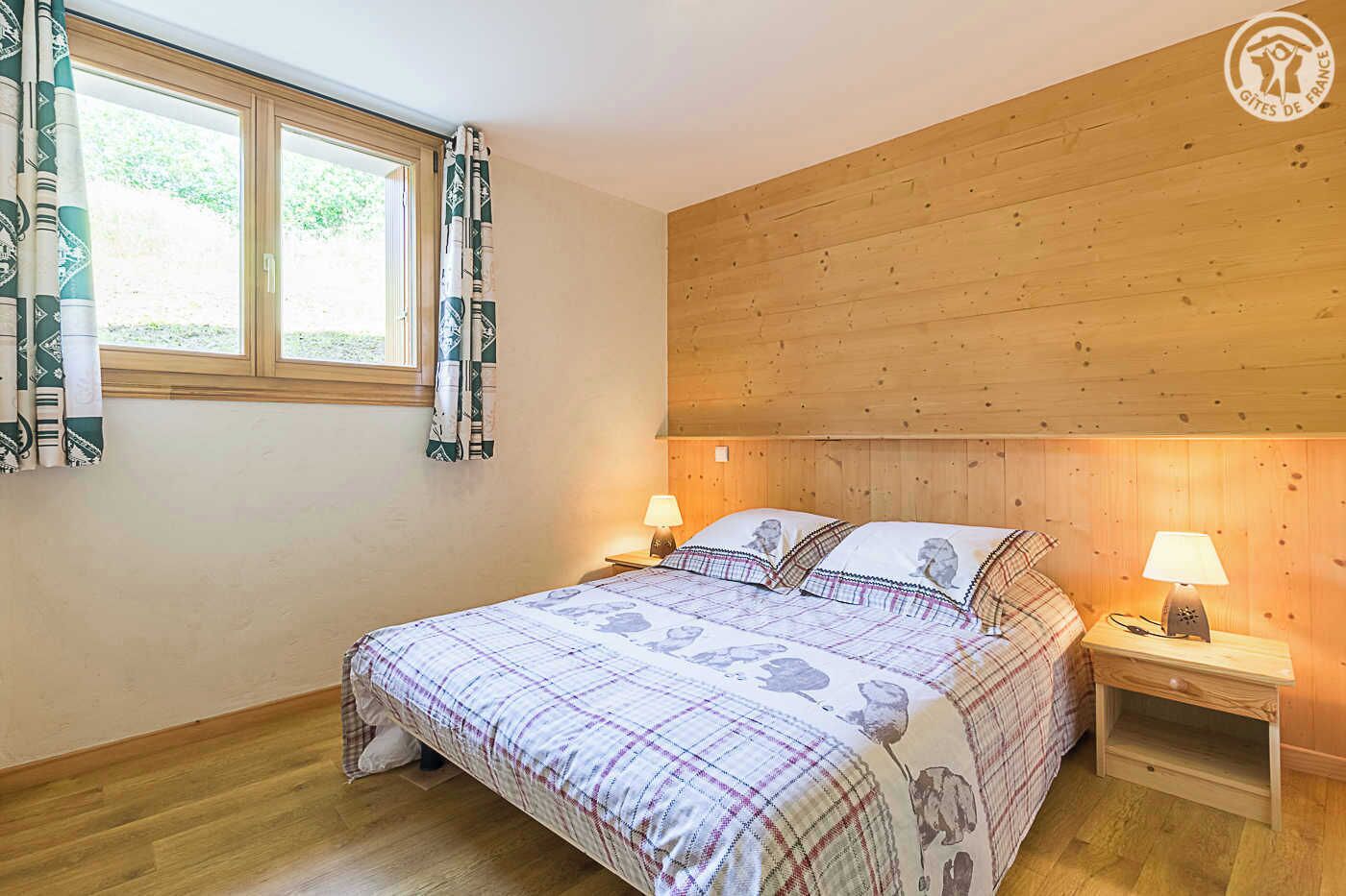 Un dormitorio con cabecera de madera, dos mesitas de noche con lámparas, cama con colcha escocesa y ventana con cortinas estampadas.
