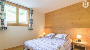 Une chambre à coucher avec une tête de lit en bois, deux tables de chevet avec des lampes, un lit recouvert d’une couverture à carreaux et une fenêtre ornée de rideaux à motifs.