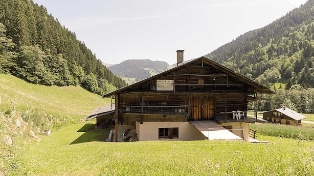 Une cabane en bois rustique, agrémentée d’une enseigne, entourée de collines verdoyantes et adossée à une montagne boisée.