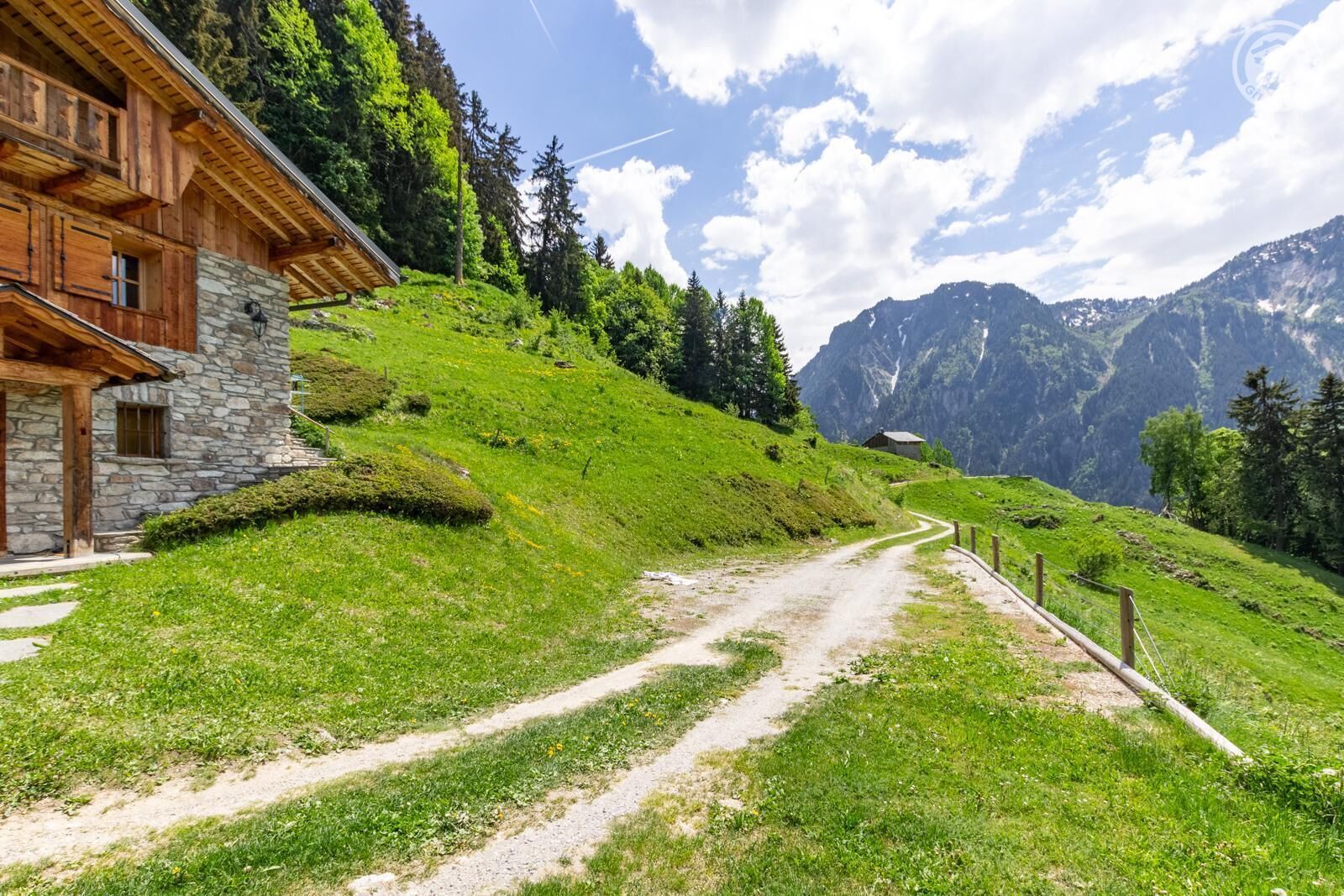 Ein Berghaus mit Steinaußenwand und Holzbalken, umgeben von üppigem Grün und einem Feldweg, der zu einer Berglandschaft führt.