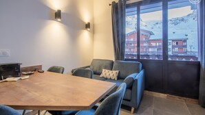 A modern dining area with a wooden table and blue chairs, a sofa with a patterned pillow, and a view of snow-covered buildings through a large window. - ROND-POINT DES PISTES - INTERLUDE (TIGNES)