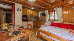 A cozy room with a wooden bed, a dining table set for two, a kitchen with wooden cabinets, and a clock on the wall. - LES COLOMBIERES - MÉSANGE (BONNEVAL SUR ARC)
