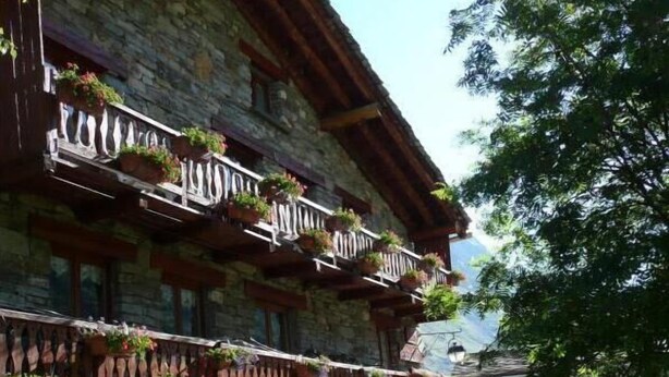 A traditional stone building with wooden balconies, outdoor seating, and lush greenery. - LES COLOMBIERES - MÉSANGE (BONNEVAL SUR ARC)
