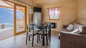 L'intérieur d'un chalet en bois comprend une table à manger, des chaises, un canapé, une cheminée et une grande fenêtre avec des rideaux.