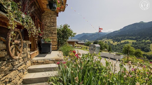 A stone building with a wooden wheel, a small outdoor seating area, and a view of a mountainous landscape. - LA FERME D'EUGENIE - FLUMET (FLUMET)