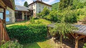 Ein rustikales Steinhaus mit überdachter Veranda und Garten, umgeben von einem Holzzaun und viel Grün.
