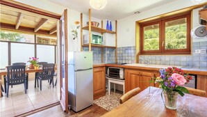 Eine kompakte Küche mit Holzausstattung, einem Kühlschrank, einer Mikrowelle und einem Spülbecken. Ein Essbereich mit Stühlen und Tisch lädt zum Verweilen ein, und ein Fenster bietet Blick ins Grüne.