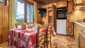 Una cuina acollidora amb armaris de fusta, una taula de dinar amb mantell vermell i blanc, i vistes a les muntanyes per les finestres.