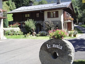 Ein Steinschild mit der Aufschrift 'Le moulin', ein Holzgebäude mit Satteldach und ein gepflegter Garten mit Blumen und Sträuchern.