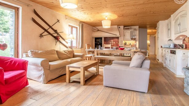 Una sala de estar acogedora con un sofá, un sillón rojo y una mesa de centro de madera. Hay una zona de cocina con armarios blancos y una mesa de comedor.