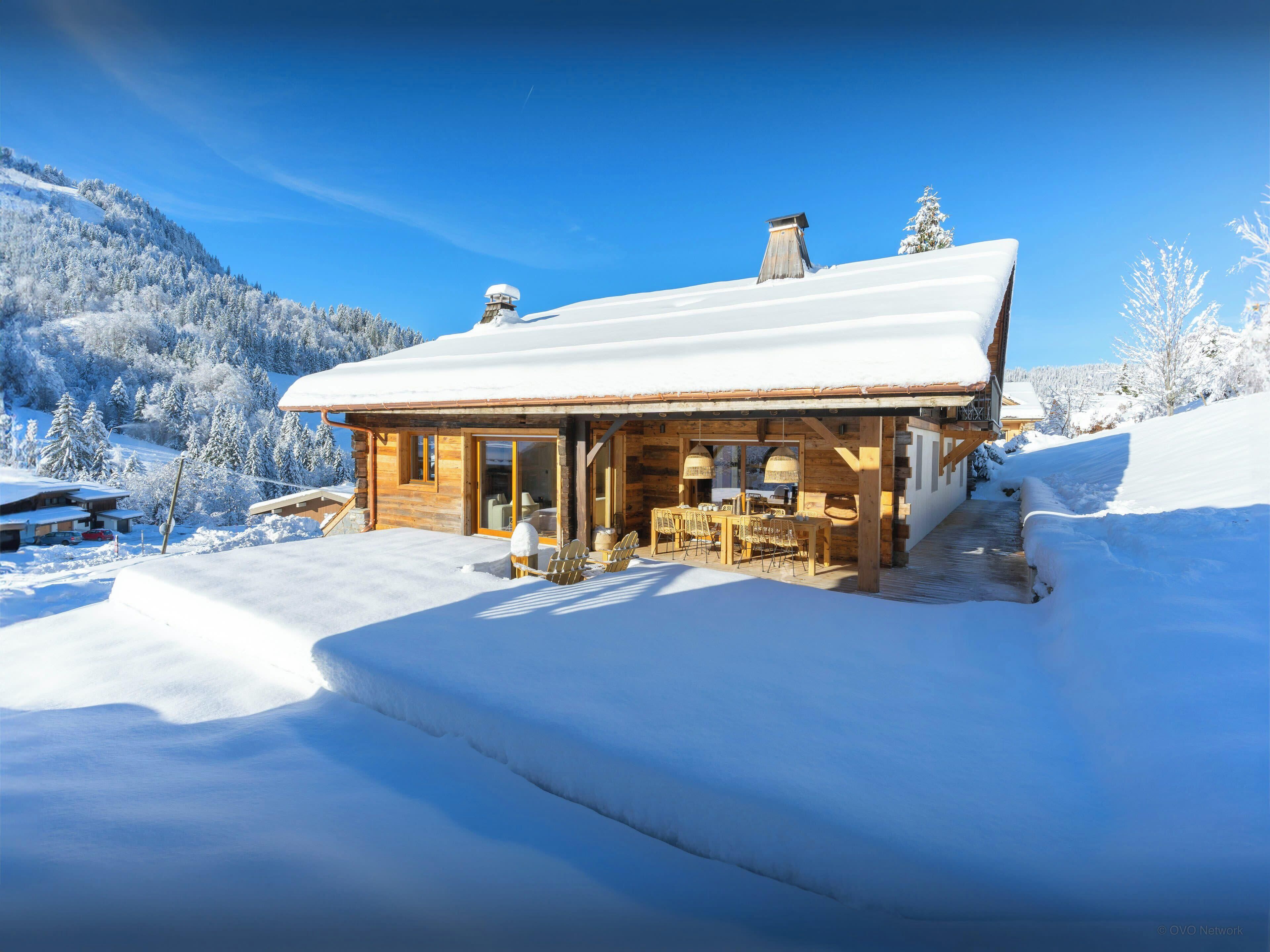 A wooden cabin with a snow-covered roof, a patio with outdoor seating, and a view of snow-covered trees and mountains.