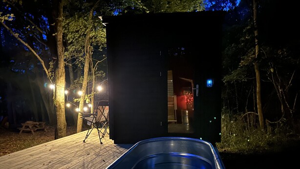 Una terraza de madera con un jacuzzi, una estructura negra pequeña y luces colgantes.