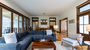 Amplia sala de estar con un sofá seccional grande, una mesa de centro de madera y un sillón de color gris claro. Se observa un comedor con sillas de madera y una mesa al fondo.