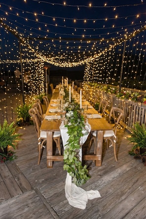 An outdoor dining area with a long wooden table set for a meal, surrounded by chairs, and decorated with string lights. - Hemingway & Mark Twain Estate | Weddings & Events | 32BR Sleeps 72 | OBX (Corolla)