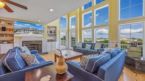 A spacious living room with a fireplace, large windows, and a view of the beach. - Hemingway & Mark Twain Estate | Weddings & Events | 32BR Sleeps 72 | OBX (Corolla)
