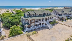 A beachfront property with a large house featuring multiple levels, a balcony, and a staircase leading to the beach. - Hemingway & Mark Twain Estate | Weddings & Events | 32BR Sleeps 72 | OBX (Corolla)