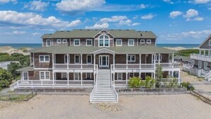 A two-story beachfront house with a front porch, a staircase leading to the entrance, and a sandy beach in the background.
