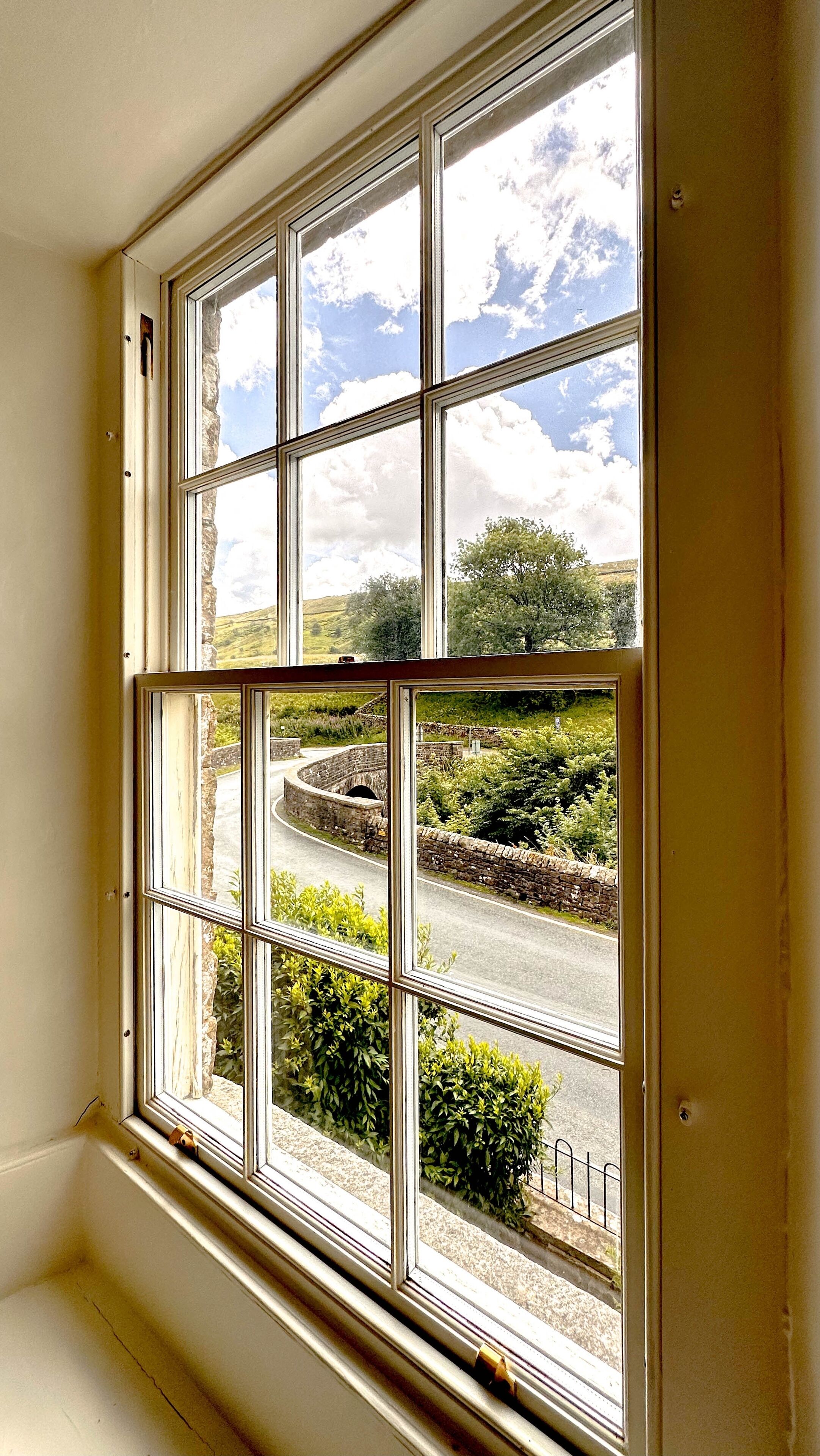 A window with a view of a stone wall, greenery, and a clear sky.