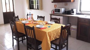Un comedor con una mesa dispuesta para cuatro, sillas de madera, una cocina con microondas y un ventanal con cortinas.