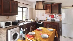Una cocina con armarios de madera, microondas, lavadora, una mesa de comedor con mantel amarillo y un refrigerador.