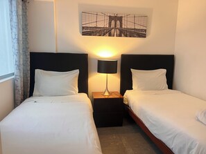 Une chambre d’hôtel avec deux lits simples, une table de chevet avec une lampe, une œuvre d’art représentant un pont accrochée au mur, et une fenêtre avec des rideaux.