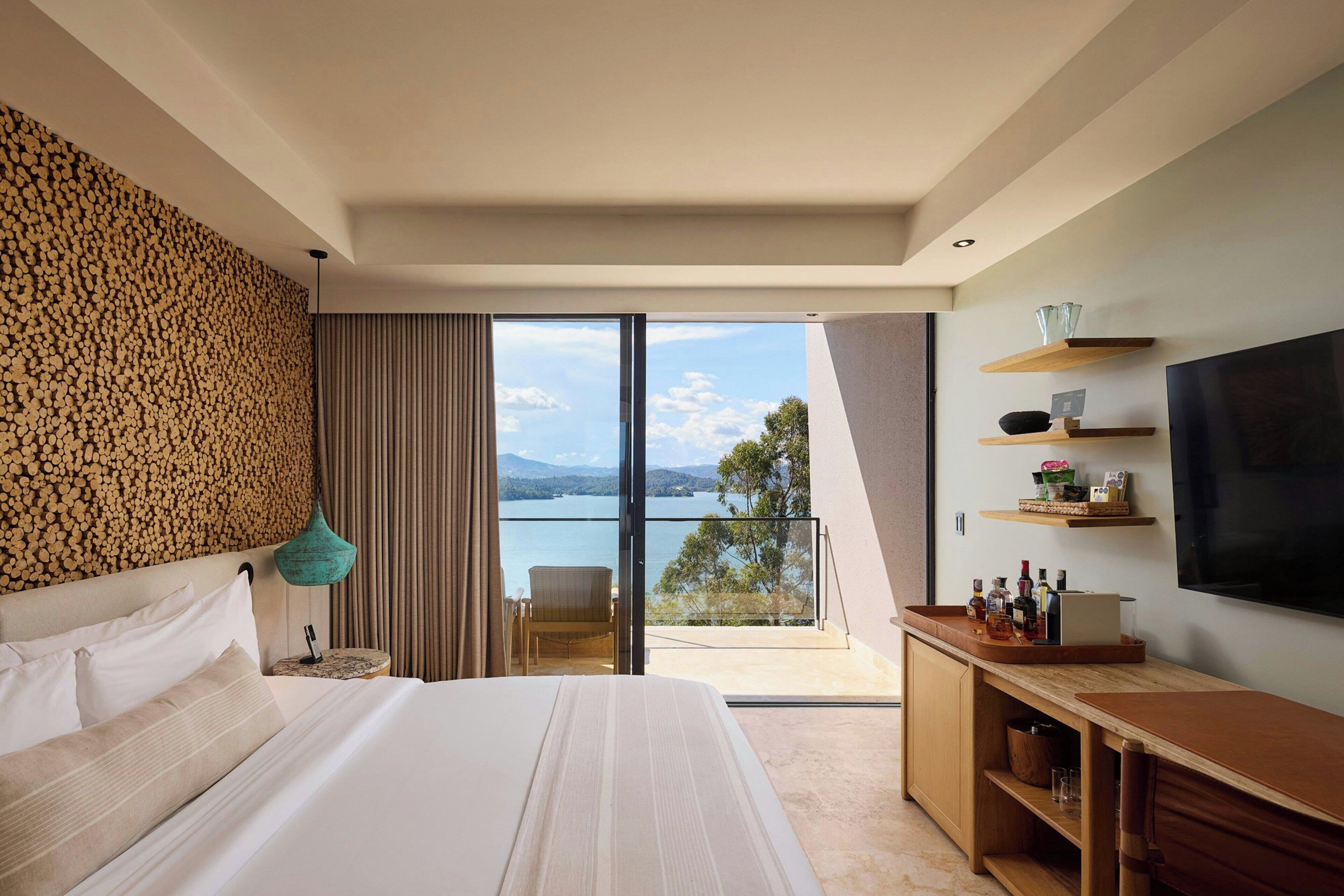 A modern hotel room with a large bed, a balcony with a view of the sea, a television, and a minibar.