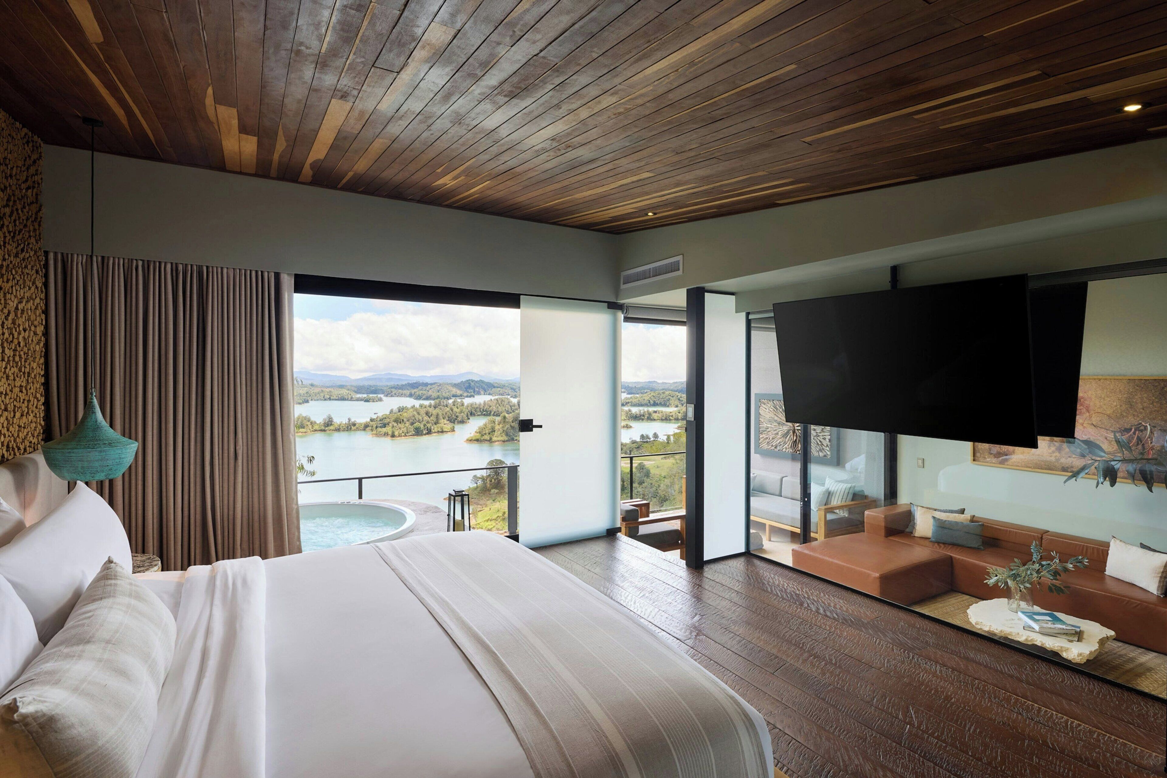 A modern bedroom with a large bed, a TV, and a view of a lake.