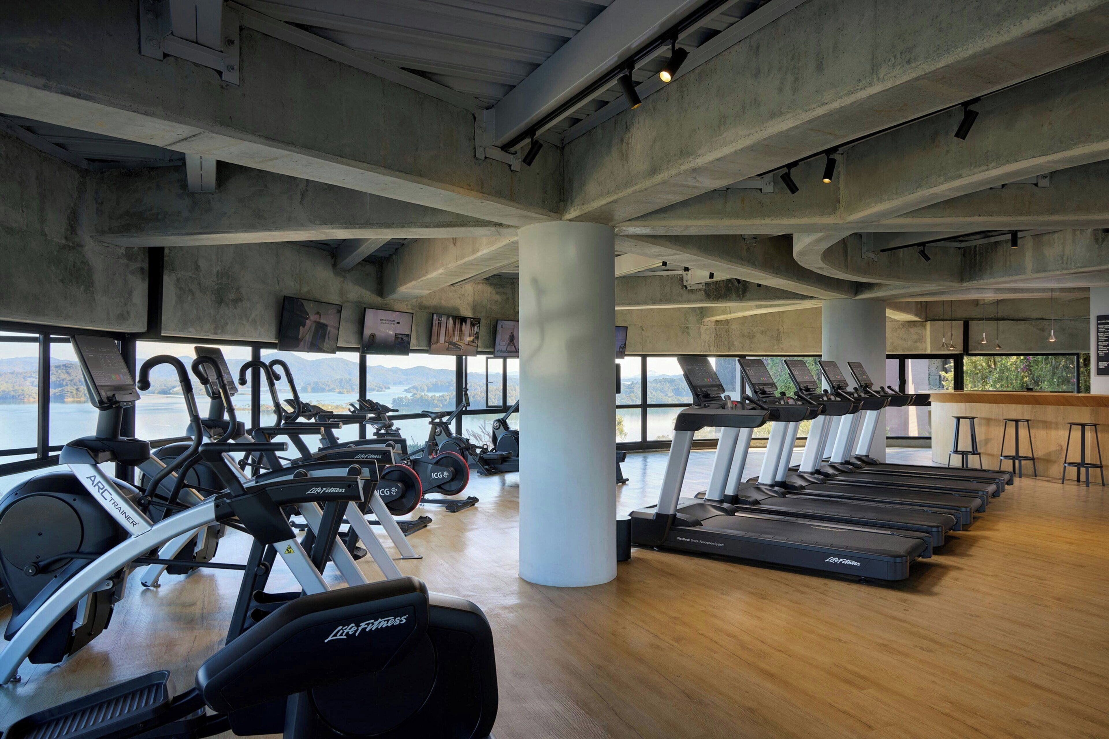 Un gimnasio con equipo cardiovascular, ventanas amplias y suelo de madera.