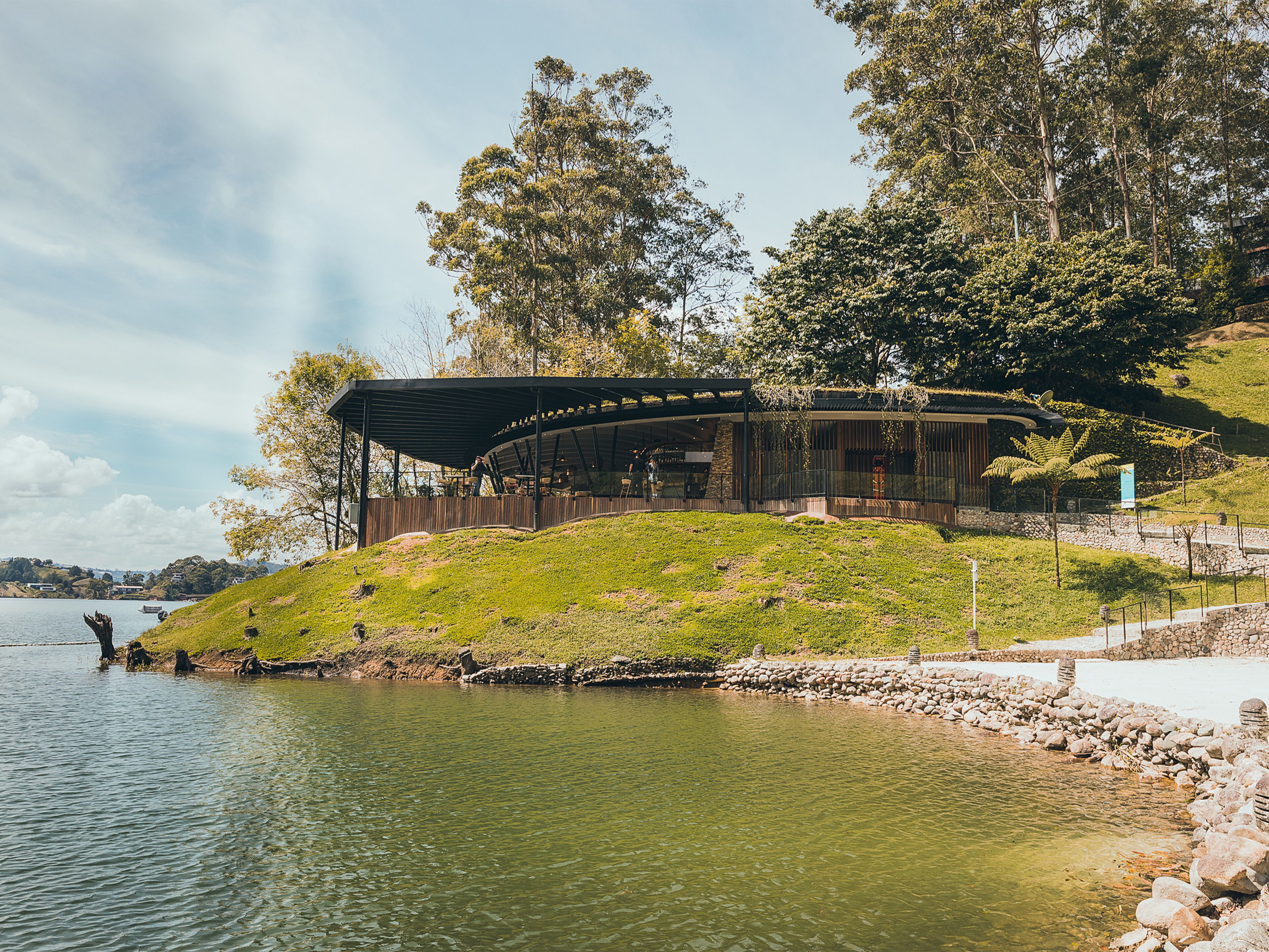 A modern building with a curved roof near a body of water, surrounded by trees and a grassy hill.