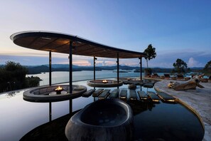 A luxurious outdoor hot tub area with a curved wooden canopy, surrounded by stone seating and a view of the water and mountains.