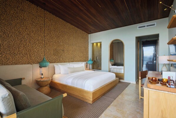 A modern hotel room with a wooden ceiling, a large bed, a sitting area with a sofa and a small table, and a bathroom visible through an archway.