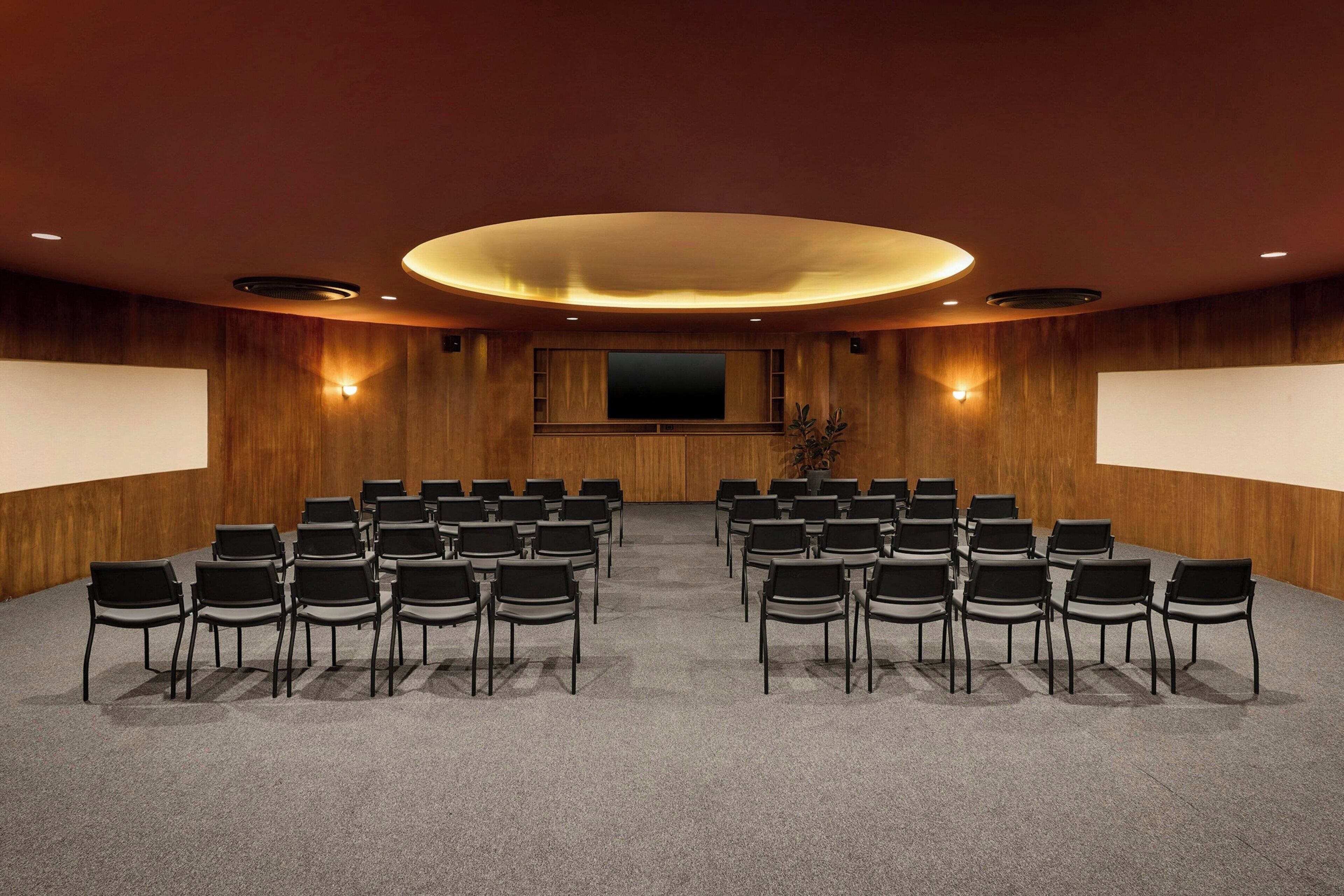 Une salle de conférence avec des rangées de chaises disposées face à un grand écran, des murs recouverts de panneaux en bois et un éclairage encastré.