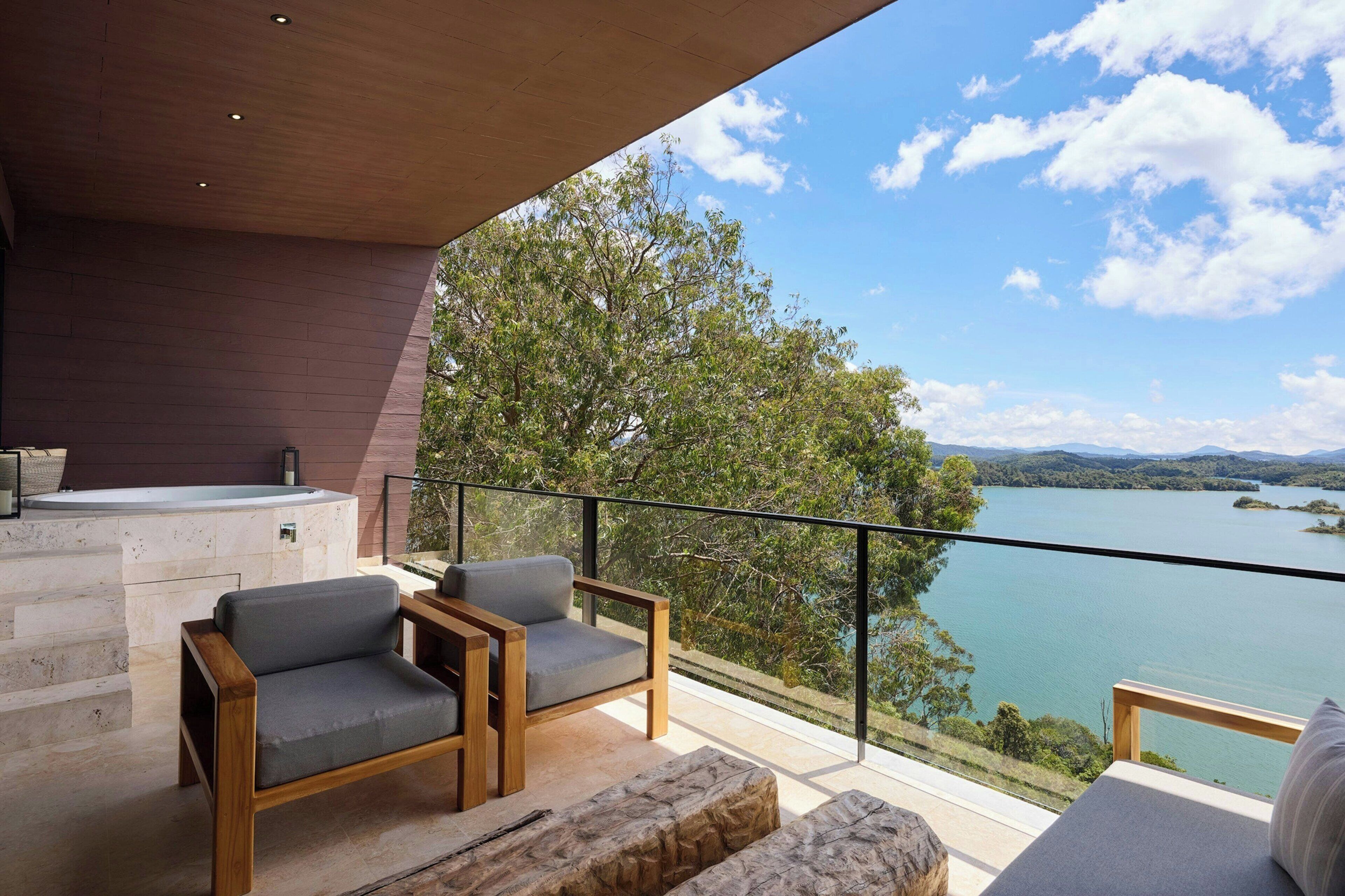 A balcony with a wooden chair, a stone bench, and a view of a lake and mountains.