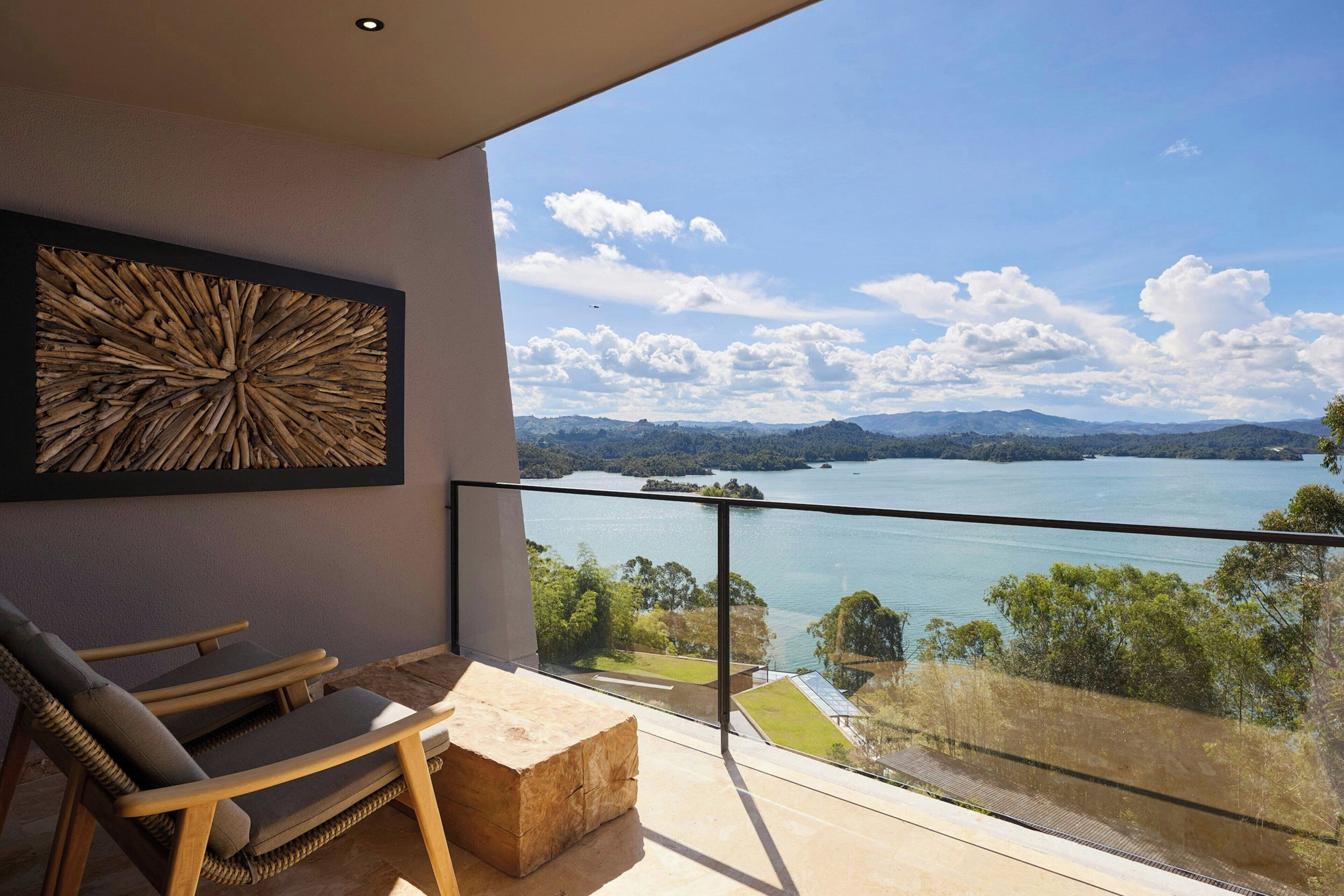 Un balcón con vistas a un lago y montañas, una silla de madera, un banco de piedra y una obra de arte colgada en la pared.