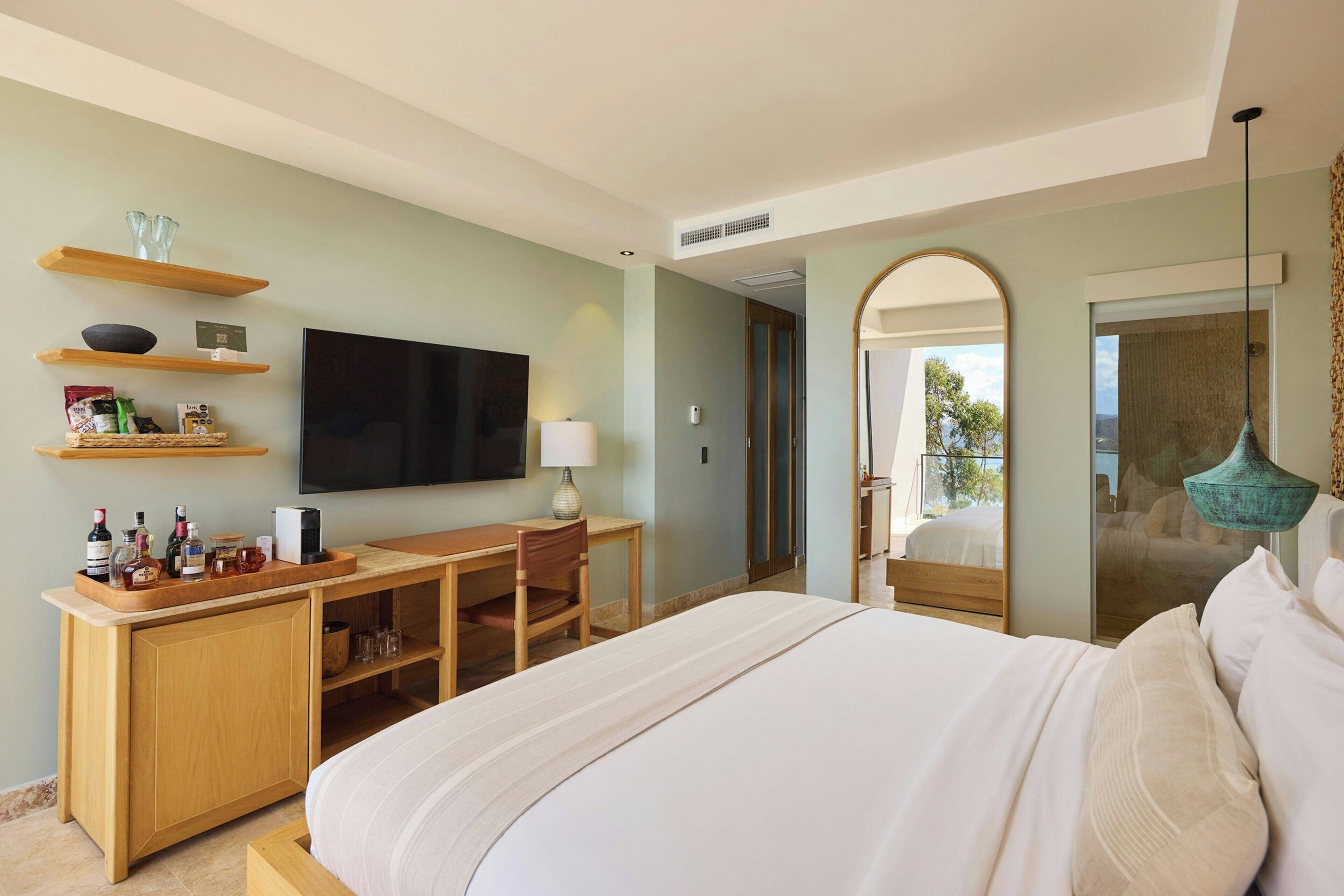 Habitación de hotel con cama, escritorio con silla, televisor y estantes con objetos.