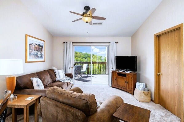 A living room with a brown leather sofa, a wooden TV stand, a ceiling fan, and a large window with a view of greenery.