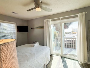 A bedroom with a bed, a ceiling fan, a TV mounted on the wall, and a view of the outside through a sliding glass door.
