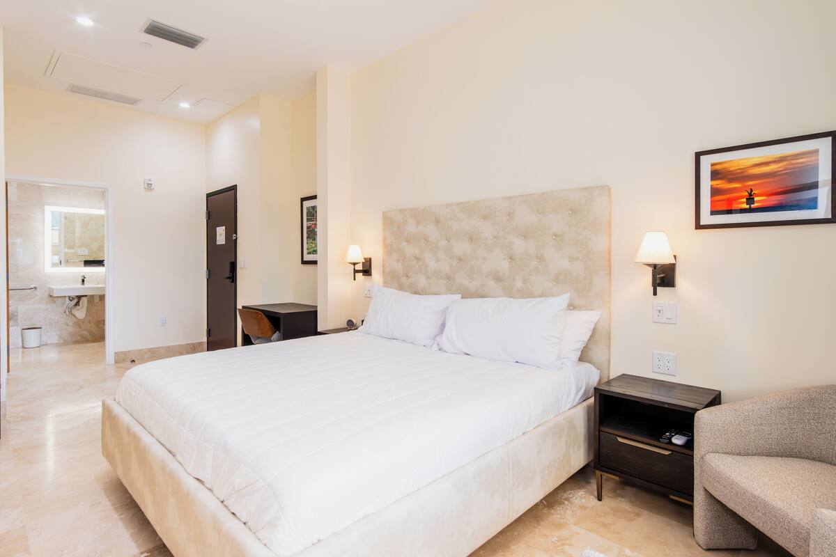 Habitación de hotel con una cama grande, una mesita de noche, una silla, un cuadro y un baño visible a través de una puerta abierta.