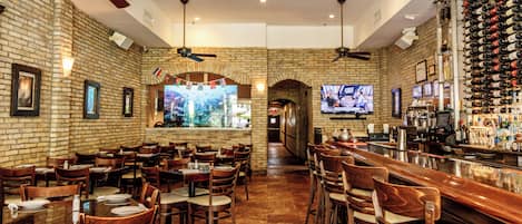 A restaurant interior with brick walls, wooden tables and chairs, ceiling fans, and a bar area with a wine rack.