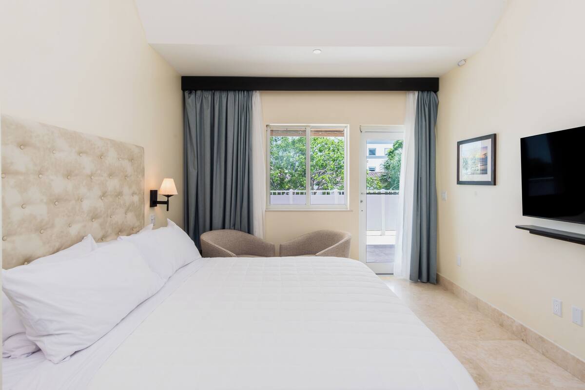 Habitación de hotel con una cama grande, televisión montada en la pared, una silla y una ventana con cortinas.