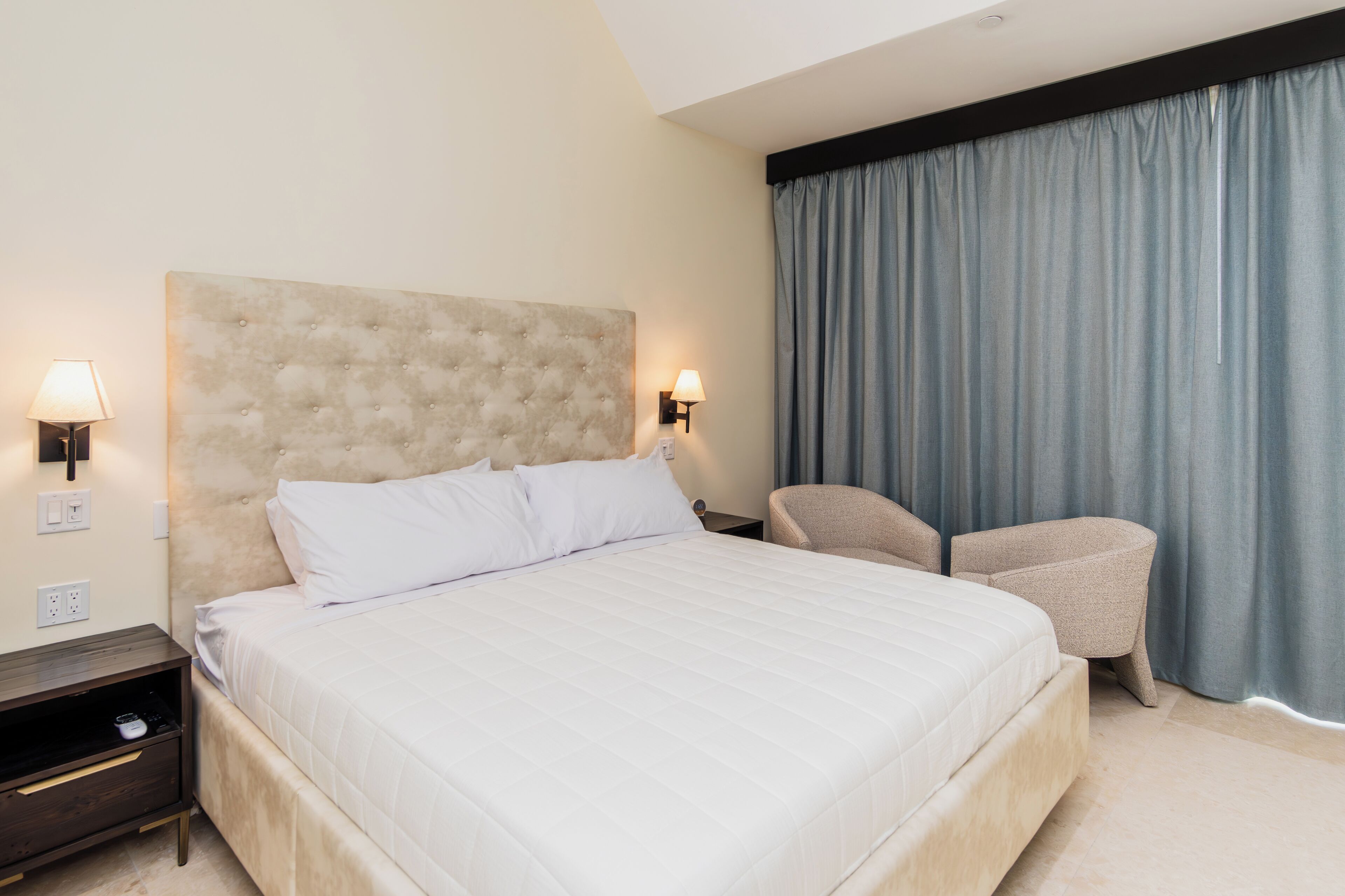 Quarto de hotel com uma cama grande, duas mesas de cabeceira, uma cadeira e uma janela com cortinas.